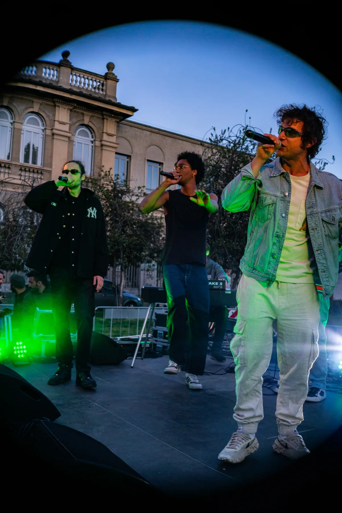 Tre voci insieme sul palco all'aperto sotto una luce verde, il palazzo storico alle spalle